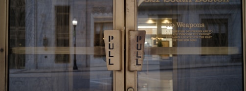 Two glass doors with large pull handles, framed by an exterior urban setting. Visible text on one door includes a 'No Weapons' policy and instructions for deliveries.