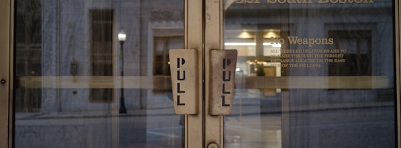 Two glass doors with large pull handles, framed by an exterior urban setting. Visible text on one door includes a 'No Weapons' policy and instructions for deliveries.