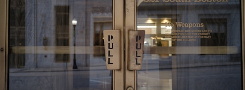 Two glass doors with large pull handles, framed by an exterior urban setting. Visible text on one door includes a 'No Weapons' policy and instructions for deliveries.