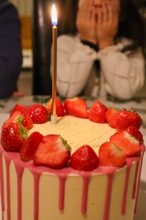 A tall cake adorned with strawberries and pink icing drips down the sides. A single lit candle stands on top of the cake. In the background, a person with long hair covers their face with their hands, suggesting surprise or anticipation.