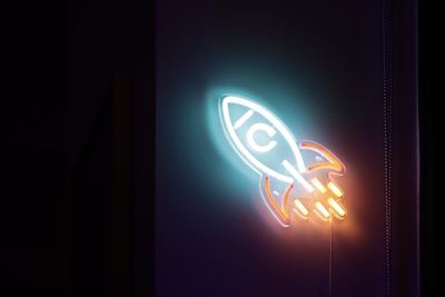 A neon sign shaped like a rocket is mounted on a dark wall, featuring glowing colors of blue, orange, and white. The central part of the rocket has a 'C' symbol, and its tail emits a vibrant orange glow, simulating flames or motion.