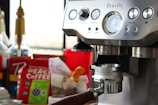 A close-up of a coffee machine with a metallic finish featuring multiple dials and buttons. In the background, there are various coffee-related items, including a bag of Peace Coffee and syrups. The scene suggests a setting for making coffee beverages.