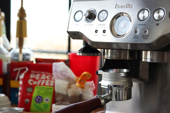 A close-up of a coffee machine with a metallic finish featuring multiple dials and buttons. In the background, there are various coffee-related items, including a bag of Peace Coffee and syrups. The scene suggests a setting for making coffee beverages.