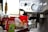 A close-up of a coffee machine with a metallic finish featuring multiple dials and buttons. In the background, there are various coffee-related items, including a bag of Peace Coffee and syrups. The scene suggests a setting for making coffee beverages.