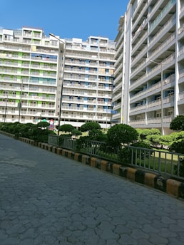 Several multi-story residential buildings with balconies, surrounded by neatly trimmed bushes and a paved walkway. The buildings have a mix of white, green, and blue colors.