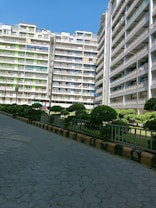Several multi-story residential buildings with balconies, surrounded by neatly trimmed bushes and a paved walkway. The buildings have a mix of white, green, and blue colors.