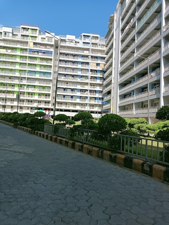 Several multi-story residential buildings with balconies, surrounded by neatly trimmed bushes and a paved walkway. The buildings have a mix of white, green, and blue colors.