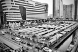 A black and white image of an urban area featuring a large, modern building with a unique design. In the foreground, there is a bustling market with rows of canopied stalls. Numerous people can be seen walking through the market, and several vehicles are parked along the adjacent streets. Tall skyscrapers are visible in the background.