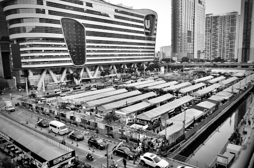 A black and white image of an urban area featuring a large, modern building with a unique design. In the foreground, there is a bustling market with rows of canopied stalls. Numerous people can be seen walking through the market, and several vehicles are parked along the adjacent streets. Tall skyscrapers are visible in the background.
