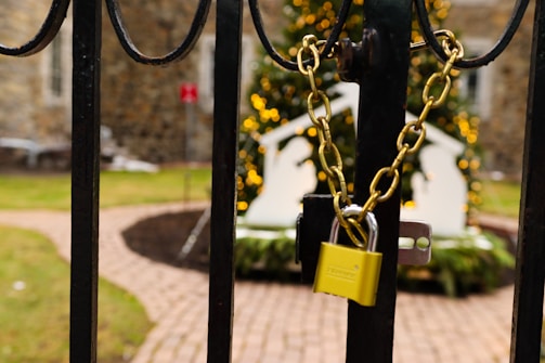 A sturdy iron gate is secured with a golden chain and padlock, with a blurred background showing a festive scene featuring warm yellow lights and a white ornamental structure, possibly part of holiday decorations. The path beyond has a brick pattern, and parts of a building are visible.