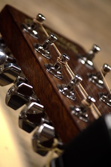 A close-up view of a guitar headstock featuring six tuning pegs made of shiny metal. The wooden texture of the headstock is visible, providing a rich brown contrast to the metallic parts. Guitar strings are wound around the tuning posts, and the background is softly blurred.