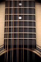 Close-up view of an acoustic guitar's strings and fretboard. The fretboard is dark with metal frets and dot inlays. The guitar body is partially visible, showcasing light-colored wood and decorative elements around the sound hole.