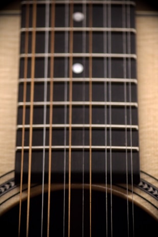 Close-up view of an acoustic guitar's strings and fretboard. The fretboard is dark with metal frets and dot inlays. The guitar body is partially visible, showcasing light-colored wood and decorative elements around the sound hole.