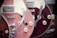 A close-up view of two electric guitars, featuring a pink and a burgundy model. The guitars have a glossy finish, with chrome hardware and distinctive retro design elements. The focus is on the body and neck areas with visible strings and control knobs.
