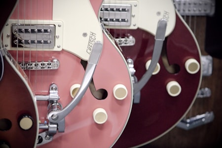 A close-up view of two electric guitars, featuring a pink and a burgundy model. The guitars have a glossy finish, with chrome hardware and distinctive retro design elements. The focus is on the body and neck areas with visible strings and control knobs.