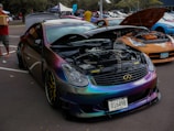 A car exhibition features a row of modified cars. A black car with a reflective, chameleon paint job is prominently displayed with its hood open, showcasing a clean engine bay. The surrounding cars are also modified, including a noticeable orange car. People are casually walking and observing the cars in an outdoor parking lot setting.
