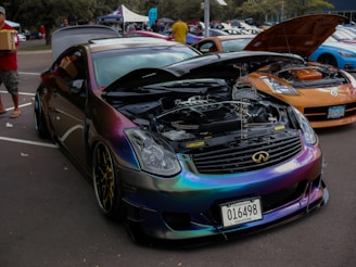 A car exhibition features a row of modified cars. A black car with a reflective, chameleon paint job is prominently displayed with its hood open, showcasing a clean engine bay. The surrounding cars are also modified, including a noticeable orange car. People are casually walking and observing the cars in an outdoor parking lot setting.