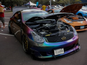 A car exhibition features a row of modified cars. A black car with a reflective, chameleon paint job is prominently displayed with its hood open, showcasing a clean engine bay. The surrounding cars are also modified, including a noticeable orange car. People are casually walking and observing the cars in an outdoor parking lot setting.