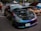 A car exhibition features a row of modified cars. A black car with a reflective, chameleon paint job is prominently displayed with its hood open, showcasing a clean engine bay. The surrounding cars are also modified, including a noticeable orange car. People are casually walking and observing the cars in an outdoor parking lot setting.