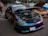 A car exhibition features a row of modified cars. A black car with a reflective, chameleon paint job is prominently displayed with its hood open, showcasing a clean engine bay. The surrounding cars are also modified, including a noticeable orange car. People are casually walking and observing the cars in an outdoor parking lot setting.