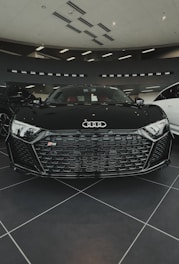 A sleek black sports car with a distinctive honeycomb grille and the Audi logo prominently displayed in a modern showroom. The interior features red seats, and the environment is well-lit with ceiling lights.