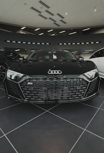 A sleek black sports car with a distinctive honeycomb grille and the Audi logo prominently displayed in a modern showroom. The interior features red seats, and the environment is well-lit with ceiling lights.