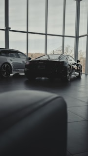 Inside a modern car showroom, two sleek luxury cars are parked. The spacious area is filled with natural light from the large floor-to-ceiling windows, casting soft shadows on the tiled floor. Trees and sky are visible through the windows, adding a serene backdrop.