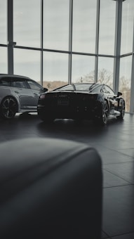 Inside a modern car showroom, two sleek luxury cars are parked. The spacious area is filled with natural light from the large floor-to-ceiling windows, casting soft shadows on the tiled floor. Trees and sky are visible through the windows, adding a serene backdrop.