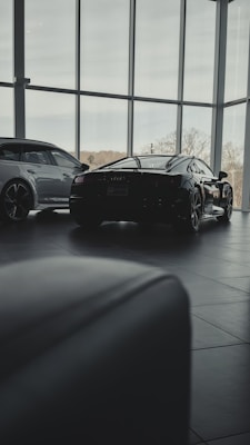 Inside a modern car showroom, two sleek luxury cars are parked. The spacious area is filled with natural light from the large floor-to-ceiling windows, casting soft shadows on the tiled floor. Trees and sky are visible through the windows, adding a serene backdrop.