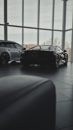 Inside a modern car showroom, two sleek luxury cars are parked. The spacious area is filled with natural light from the large floor-to-ceiling windows, casting soft shadows on the tiled floor. Trees and sky are visible through the windows, adding a serene backdrop.