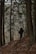 A person with a large backpack walks along a narrow, forested hiking trail. Tall trees with a dense canopy line the path, and brown leaves cover the ground, indicating a late autumn or early winter setting.
