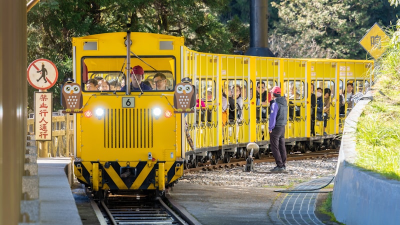 A bright yellow sightseeing train is stopped at a platform with passengers inside, surrounded by greenery. A conductor in a red cap stands beside the train, while several people look on from inside. The train features owl decorations on the front, and there are warning signs nearby.