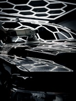 A power tool is being used for polishing a car's surface, with a geometric light pattern reflecting over the shiny black surface. The tool is white and held by a hand, emphasizing detail and precision in the task.
