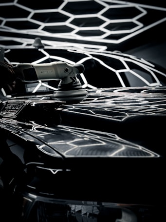 A power tool is being used for polishing a car's surface, with a geometric light pattern reflecting over the shiny black surface. The tool is white and held by a hand, emphasizing detail and precision in the task.