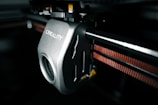 A close-up view of a 3D printer component featuring a mechanical part with the brand name 'Creality' inscribed on it. The metallic surface has a smooth finish, and there is a visible gear mechanism. An orange belt is running parallel to a metal rod, indicating functional connectivity within the printer.