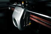 A close-up view of a 3D printer component featuring a mechanical part with the brand name 'Creality' inscribed on it. The metallic surface has a smooth finish, and there is a visible gear mechanism. An orange belt is running parallel to a metal rod, indicating functional connectivity within the printer.