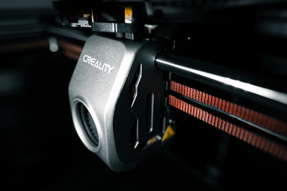 A close-up view of a 3D printer component featuring a mechanical part with the brand name 'Creality' inscribed on it. The metallic surface has a smooth finish, and there is a visible gear mechanism. An orange belt is running parallel to a metal rod, indicating functional connectivity within the printer.