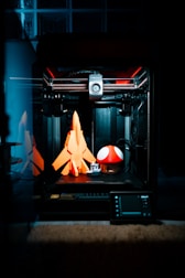 A 3D printer is in action, crafting three-dimensional objects. On the print bed are a bright orange rocket, a small white and grey boat, and a large red mushroom with white spots, reminiscent of a well-known video game item. The interior of the printer is illuminated, revealing its mechanical components and creating a focused, technological ambiance.