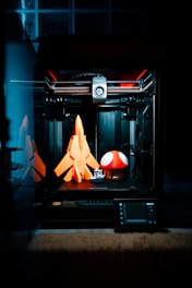 A 3D printer is in action, crafting three-dimensional objects. On the print bed are a bright orange rocket, a small white and grey boat, and a large red mushroom with white spots, reminiscent of a well-known video game item. The interior of the printer is illuminated, revealing its mechanical components and creating a focused, technological ambiance.