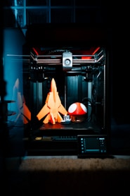 A 3D printer is in action, crafting three-dimensional objects. On the print bed are a bright orange rocket, a small white and grey boat, and a large red mushroom with white spots, reminiscent of a well-known video game item. The interior of the printer is illuminated, revealing its mechanical components and creating a focused, technological ambiance.