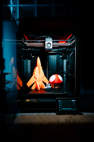A 3D printer is in action, crafting three-dimensional objects. On the print bed are a bright orange rocket, a small white and grey boat, and a large red mushroom with white spots, reminiscent of a well-known video game item. The interior of the printer is illuminated, revealing its mechanical components and creating a focused, technological ambiance.