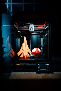 A 3D printer is in action, crafting three-dimensional objects. On the print bed are a bright orange rocket, a small white and grey boat, and a large red mushroom with white spots, reminiscent of a well-known video game item. The interior of the printer is illuminated, revealing its mechanical components and creating a focused, technological ambiance.