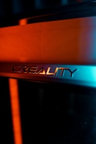 The image features the logo of Creality, likely on a 3D printer or similar device. The logo is set against a background with contrasting light, creating a dramatic effect. The lighting highlights the metallic texture of the logo, with a blend of warm orange and cool blue tones.