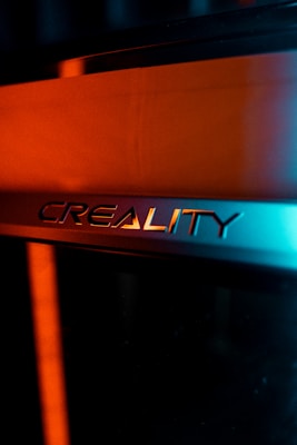 The image features the logo of Creality, likely on a 3D printer or similar device. The logo is set against a background with contrasting light, creating a dramatic effect. The lighting highlights the metallic texture of the logo, with a blend of warm orange and cool blue tones.