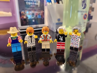 Five LEGO minifigures are displayed in a line on a reflective surface. Each has distinct outfits and accessories, such as a cowboy hat, glasses, and varied patterned clothing. The background is slightly blurred, showing part of a bottle and unclear signage.