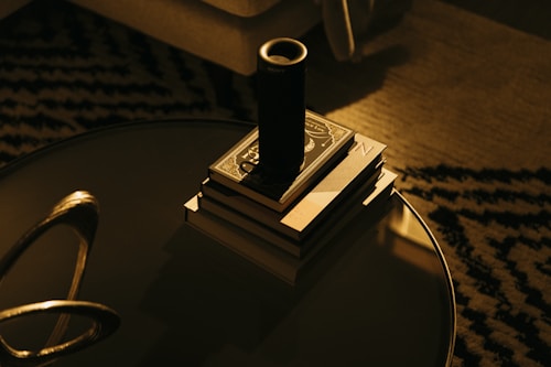 A cozy living room setting with a stack of books placed neatly on a round, reflective coffee table. A cylindrical black speaker or container is positioned on top of the books. The surface of the table is glossy, reflecting some light. The background shows a patterned carpet and part of a couch, all bathed in warm, subdued lighting.