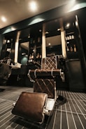A vintage-style barber chair with brown leather upholstery and chrome accents is centered in a modern barbershop setting. The room features dark wooden cabinetry with various grooming products and a large mirror. Overhead lighting casts a warm glow, highlighting the sleek design of the chair and the clean lines of the interior.