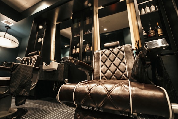 Luxurious leather barber chair with diamond stitching is prominently positioned in a stylish, modern barbershop. The background features dark shelves stocked with grooming products and bottles. A sleek sink for washing hair is visible near the chair. The lighting creates a warm, inviting atmosphere with a large ceiling lamp adding to the ambiance.
