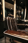 A luxurious barber chair with dark brown leather upholstery featuring white piping, set in an upscale barbershop. The chair is positioned next to shelves containing various bottles, likely grooming products or refreshments. The shop has a modern and elegant interior design, with dark colors and subtle lighting contributing to an overall sophisticated atmosphere.