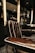 A luxurious barber chair with dark brown leather upholstery featuring white piping, set in an upscale barbershop. The chair is positioned next to shelves containing various bottles, likely grooming products or refreshments. The shop has a modern and elegant interior design, with dark colors and subtle lighting contributing to an overall sophisticated atmosphere.